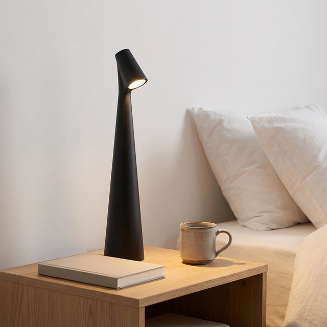 Lámpara Faro encendida sobre un velador de madera clara junto a una taza de cerámica y un libro cerrado